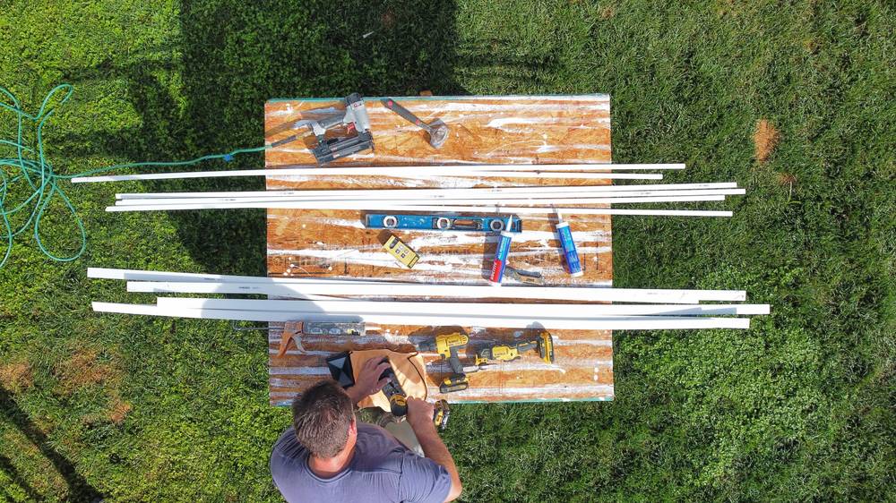 Seen from directly above: a brad nailer, hammer, spirit level, tape measure, caulk tubes, a cordless drill and a reciprocating saw laid out on a sheet of plywood in the grass, with lengths of white trim ranged either side.