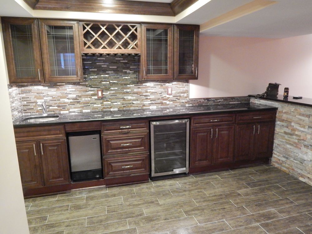 The same wet bar seen wide, showing large format wood look plank tile on the floor and a dry stacked stone veneer half wall with a dark counter cap.