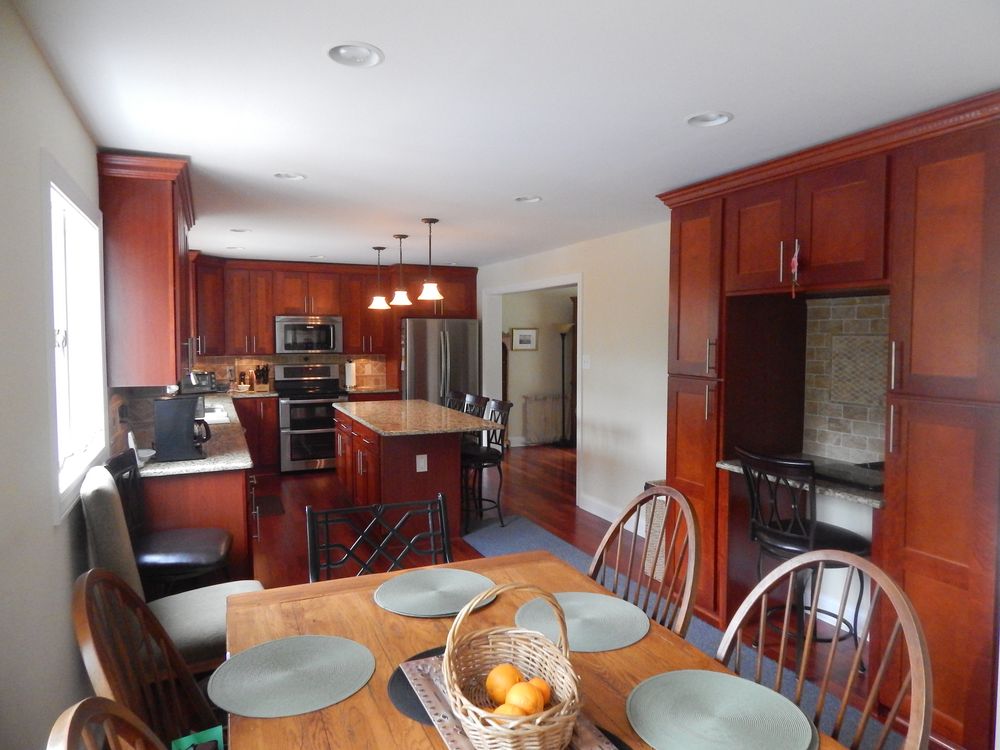 A finished cherry stained kitchen with crown moulding, a granite topped island under three pendant lights, stainless appliances, a tile backsplash and cherry hardwood floors, seen past a dining table.