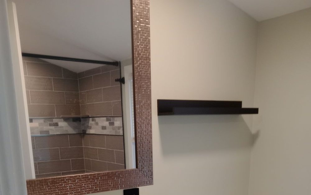 A bathroom mirror framed in copper toned mosaic tile, reflecting a walk in shower lined in large grey subway tile with a horizontal mosaic accent band and a recessed niche.