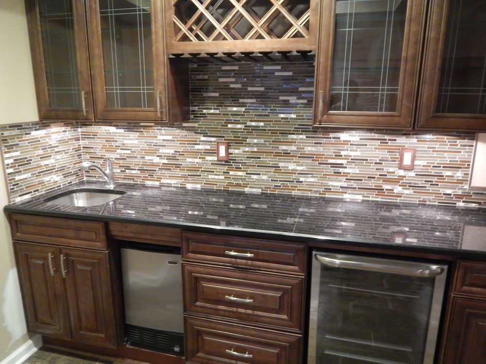 A wet bar with a linear glass and stone mosaic backsplash in cream, bronze and slate, dark speckled granite counter, stainless undermount sink and dark stained cabinets with a lattice wine rack above.